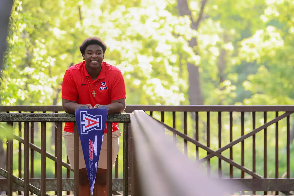 Graduate in a red shirt holding an Arizona pennant on a park bridge