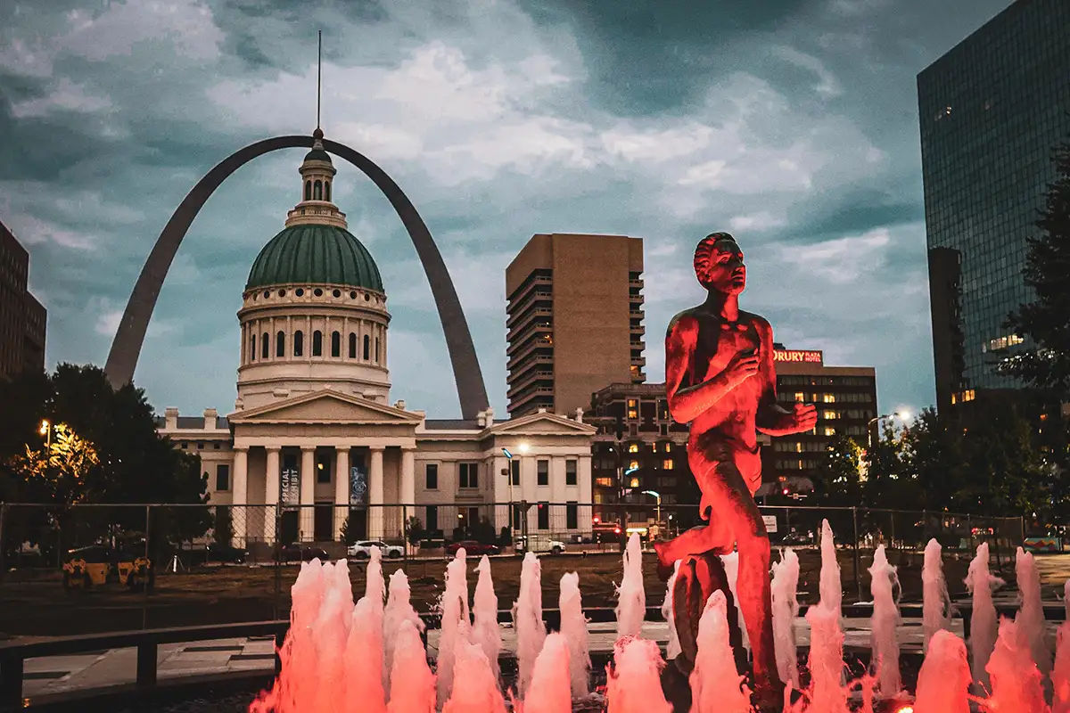 Old Courthouse and the Gateway Arch behind a statue lit red over fountains